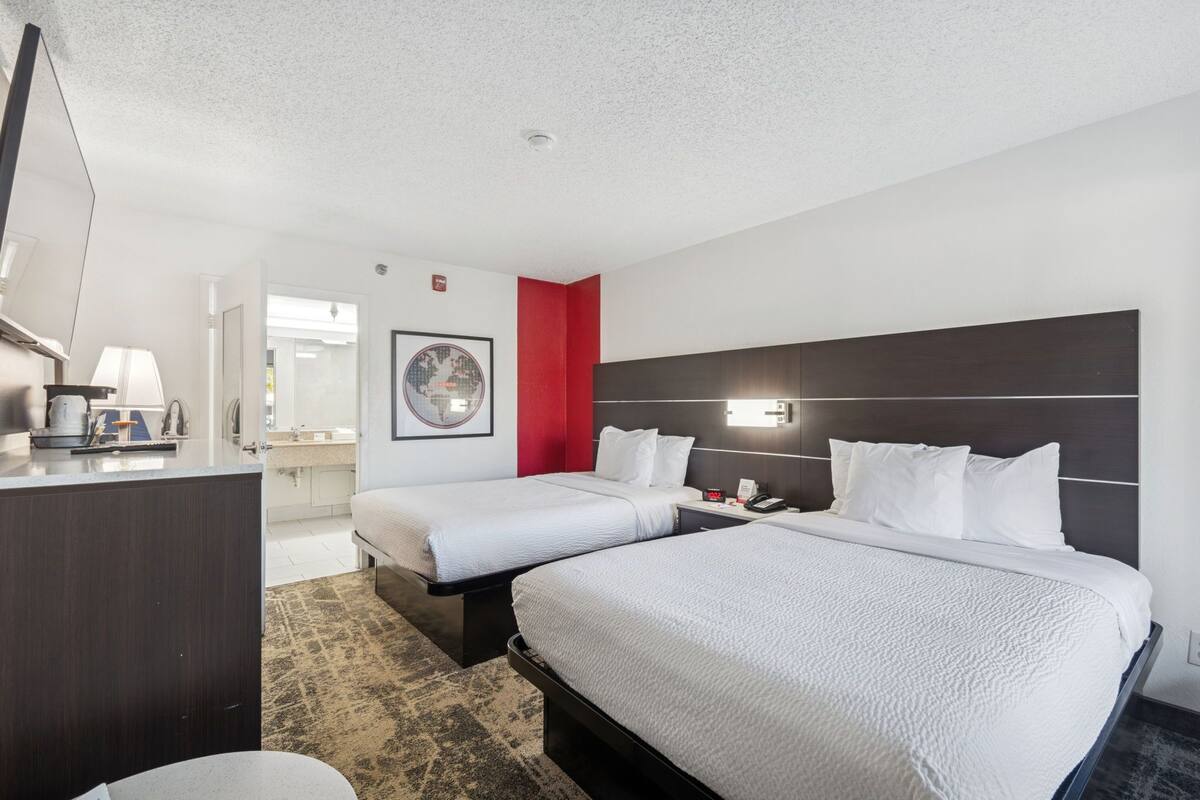 A hotel room with two beds, a desk, a mirror, a nightstand, and a bathroom visible through an open door.