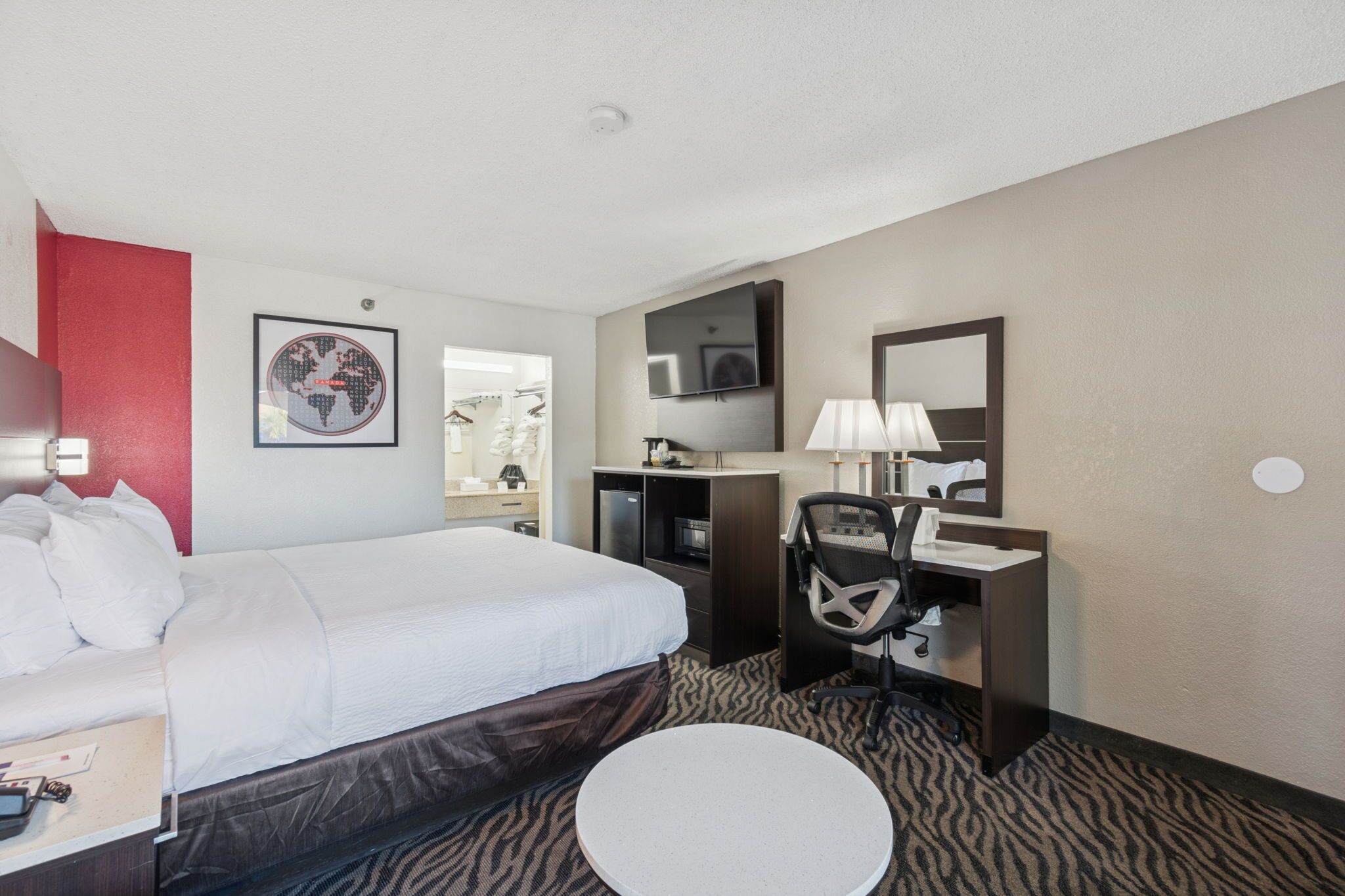 Habitación de hotel con cama, escritorio, silla, mesa redonda, televisor y espejo.
