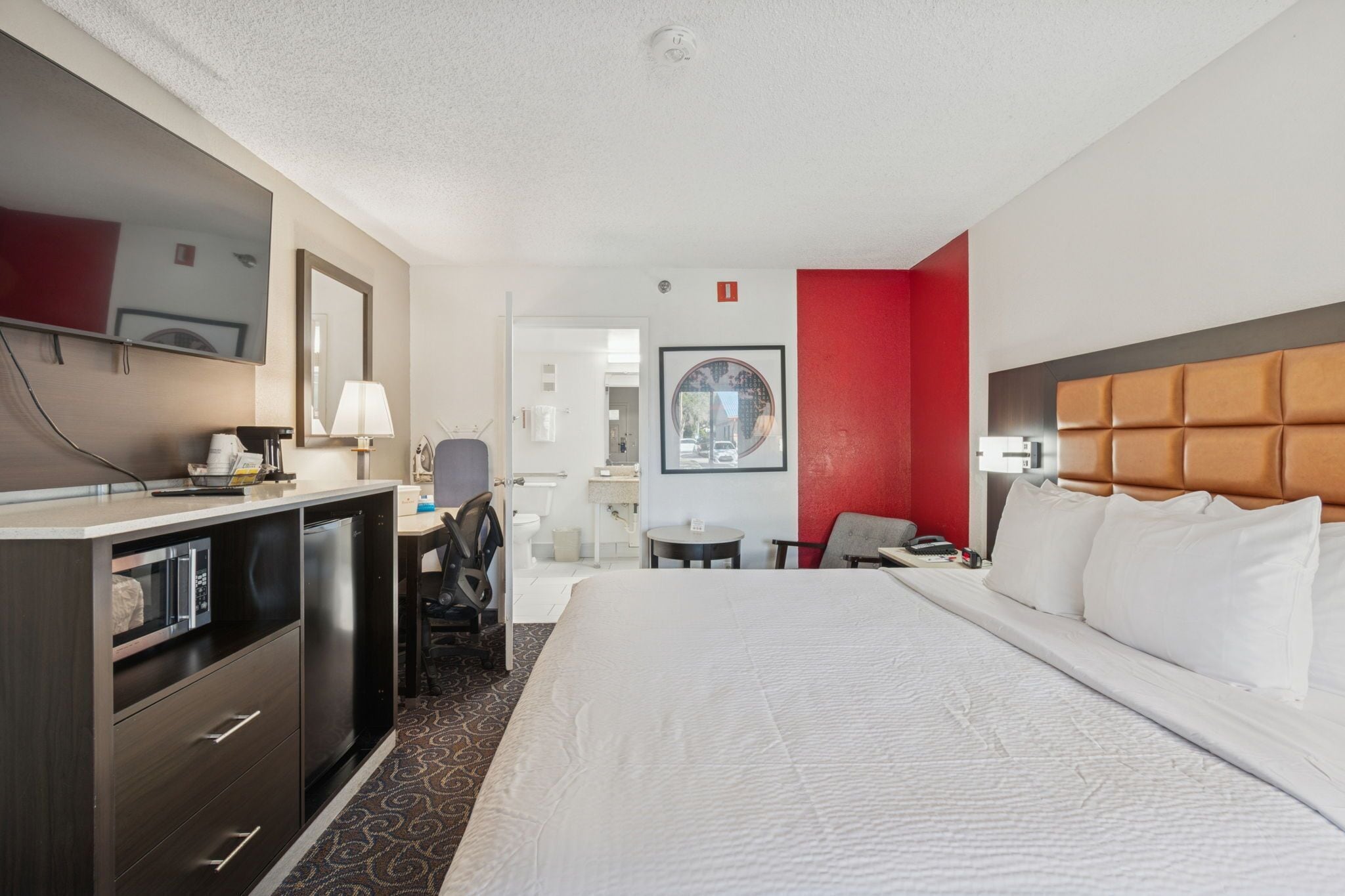 A hotel room with a large bed, a desk with a chair, a small table, a TV, and a mirror.