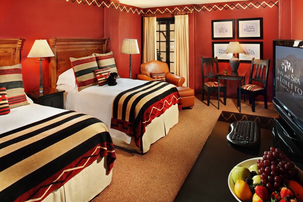 A hotel room with two beds, a desk, a chair, a television, and a fruit bowl.