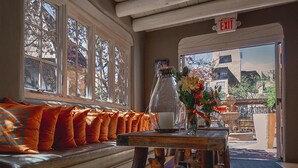 A wooden bench with orange cushions, a wooden table with a vase of flowers, and a view of the outside through large windows.