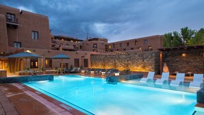 A swimming pool with lounge chairs and umbrellas, surrounded by buildings with balconies and a stone wall.