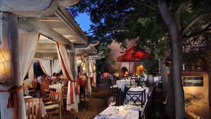 An outdoor dining area with tables set for two, wooden chairs, and a red umbrella.