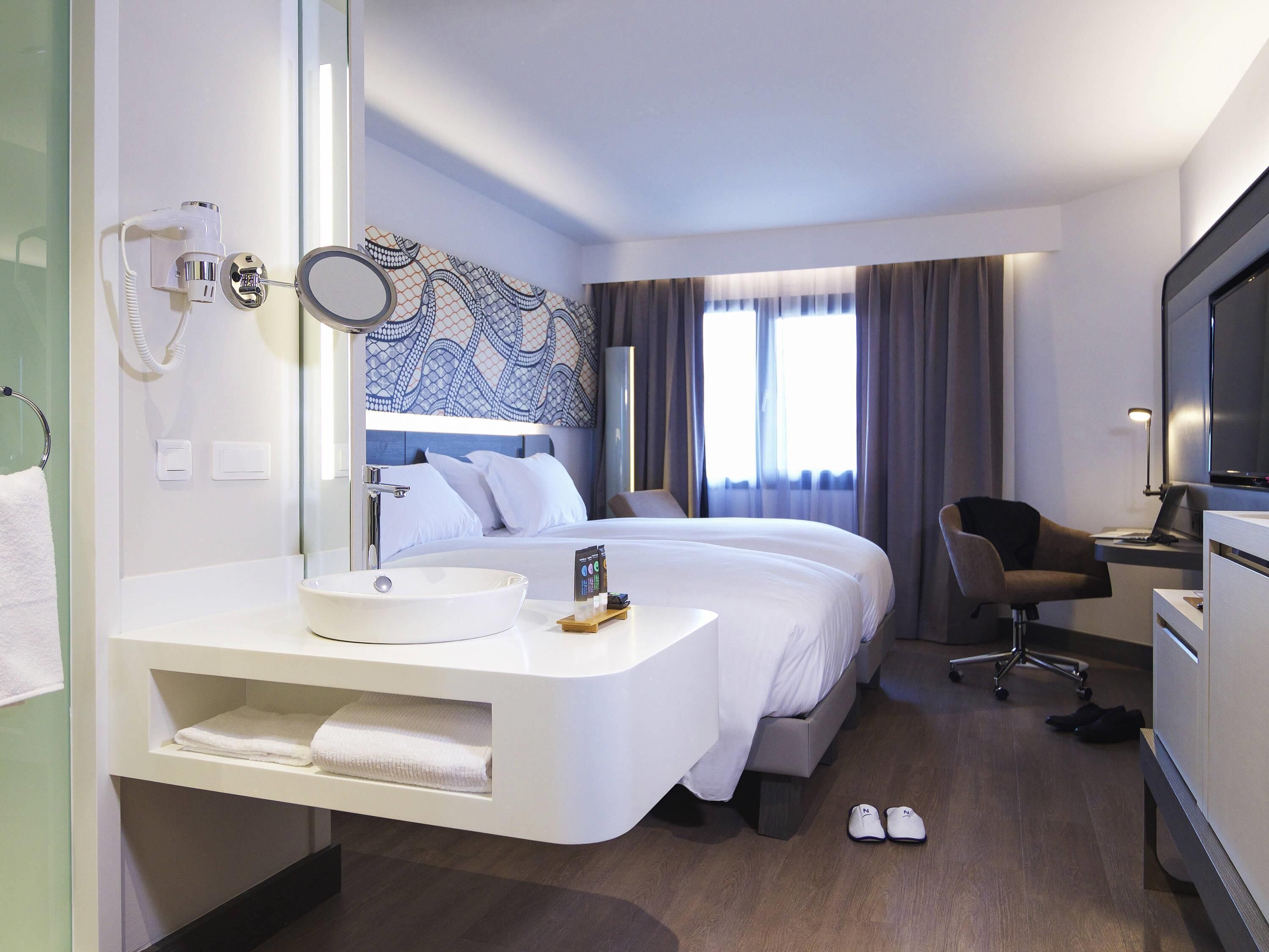 A hotel room with a bed, a desk with a computer, a bathroom with a sink, and a large mirror.