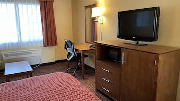 Quarto de hotel com cama, escrivaninha, cadeira, televisão, guarda-roupa e janela com cortinas.