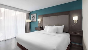 A hotel room with a large bed, a nightstand with a lamp, a wall-mounted clock, and a window with curtains. - Wingate by Wyndham Kansas City (Kansas City)