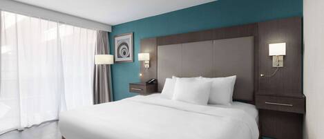 A hotel room with a large bed, a nightstand with a lamp, a wall-mounted clock, and a window with curtains.