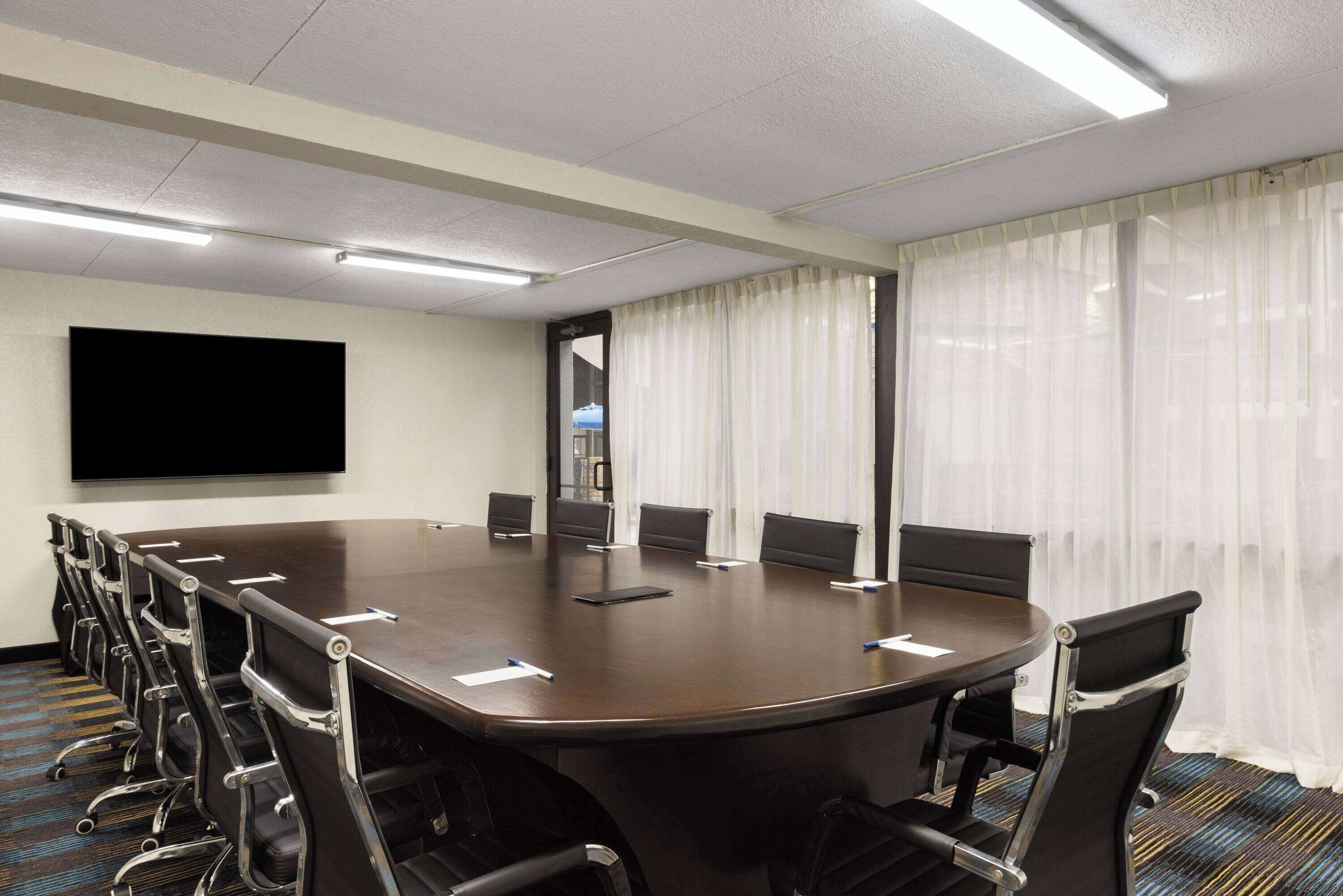 Un salón de conferencias con una larga mesa, sillas, un televisor de pantalla plana y una ventana con cortinas translúcidas.
