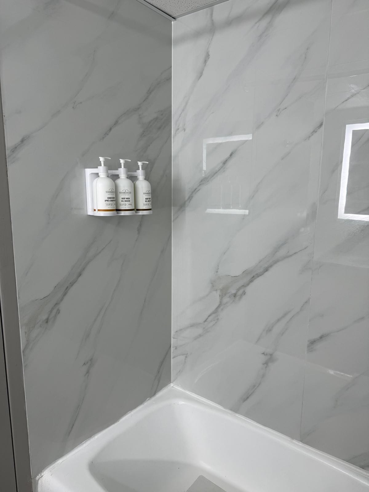 A bathroom with a marble wall, a shower area, and a shelf with three bottles of toiletries.