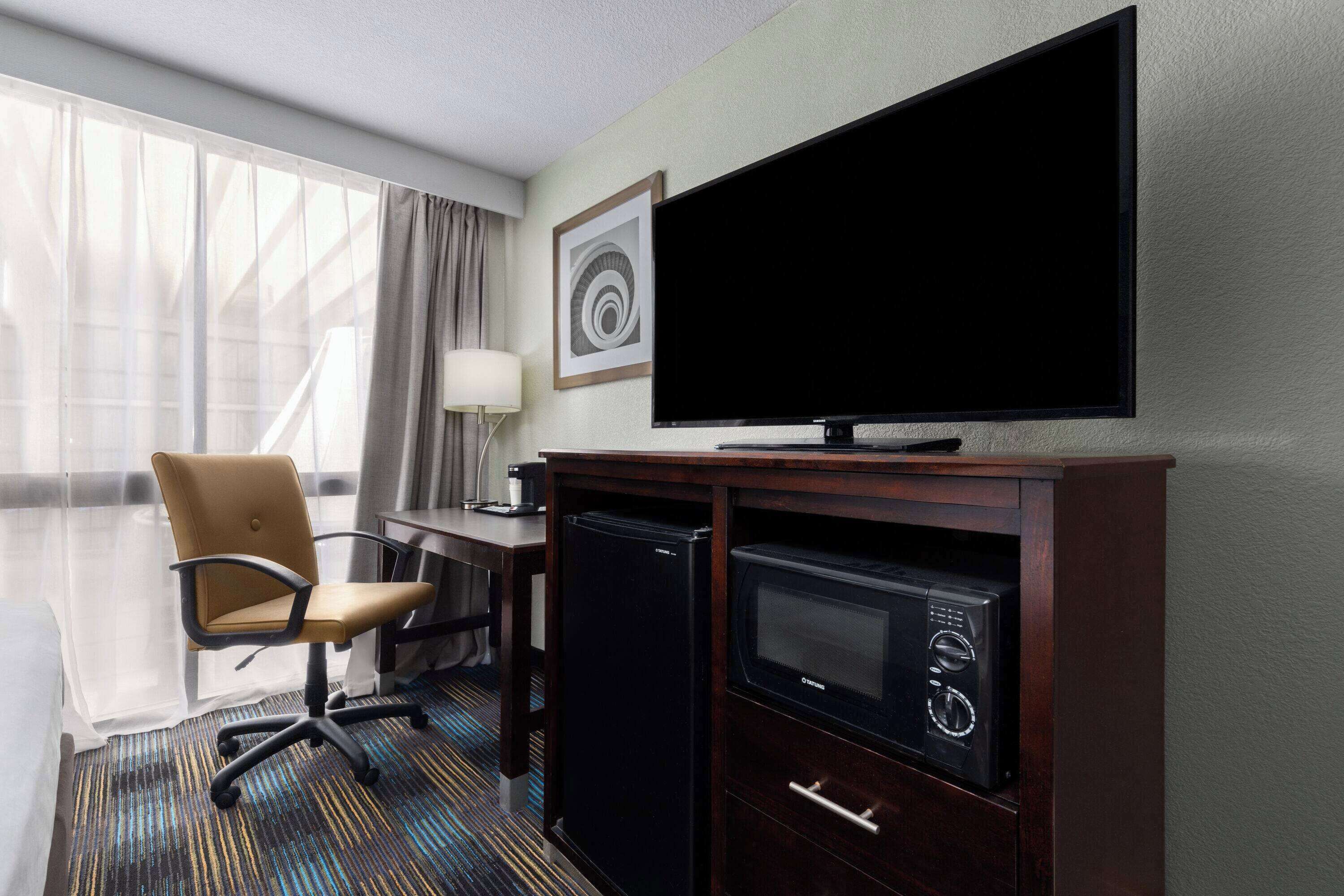 A hotel room with a large flat-screen TV, a desk with a lamp, a chair, a minibar, and a large window with sheer curtains.