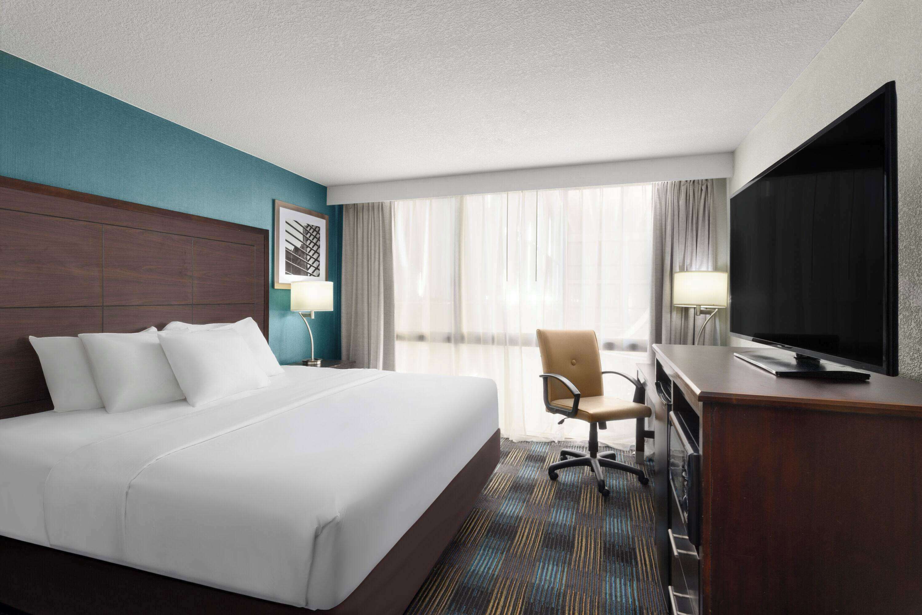 A hotel room with a large bed, a desk with a television, a chair, and a window with curtains.
