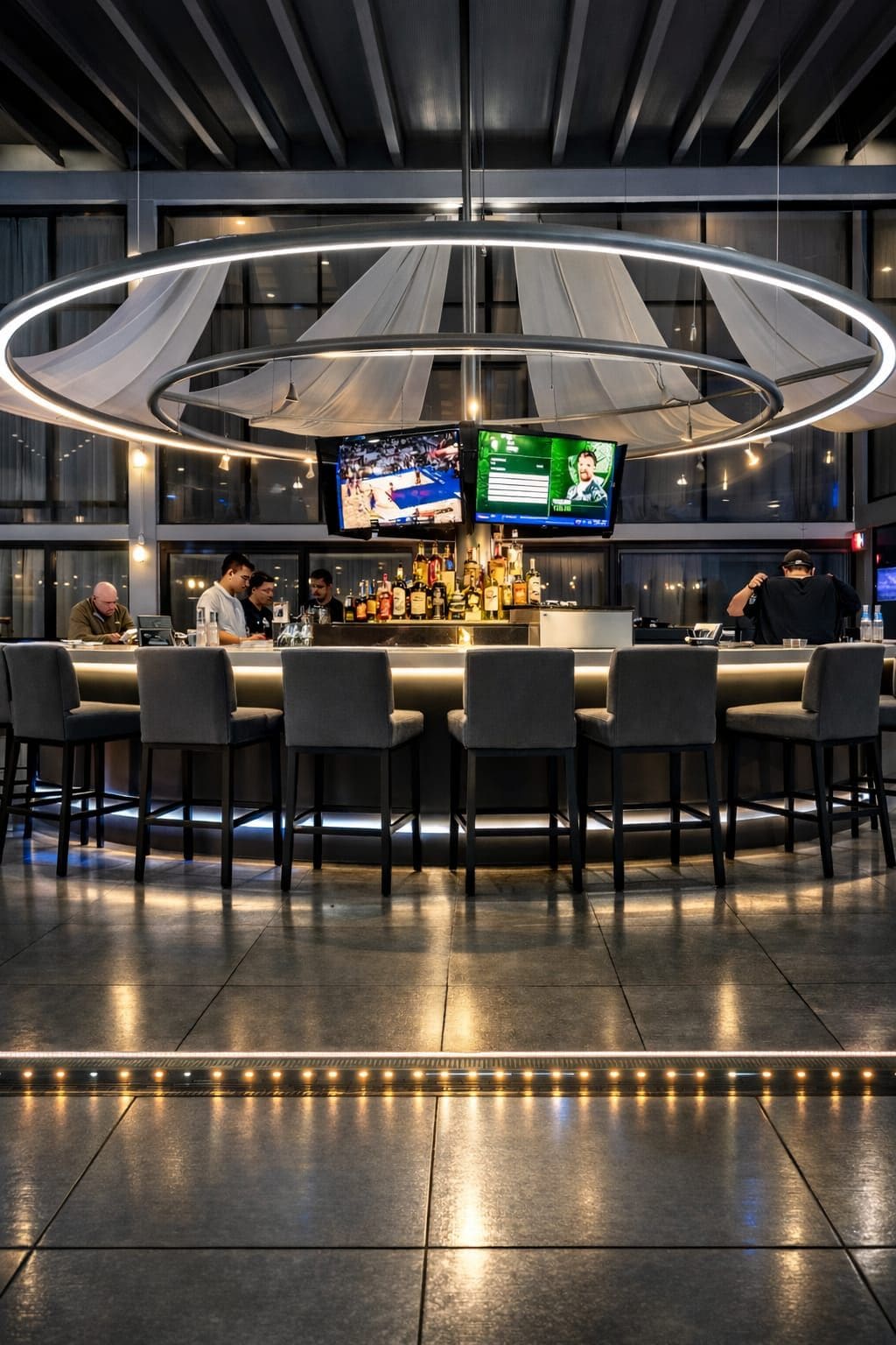 A modern bar area with multiple screens, a well-stocked bar, and a circular ceiling light fixture.