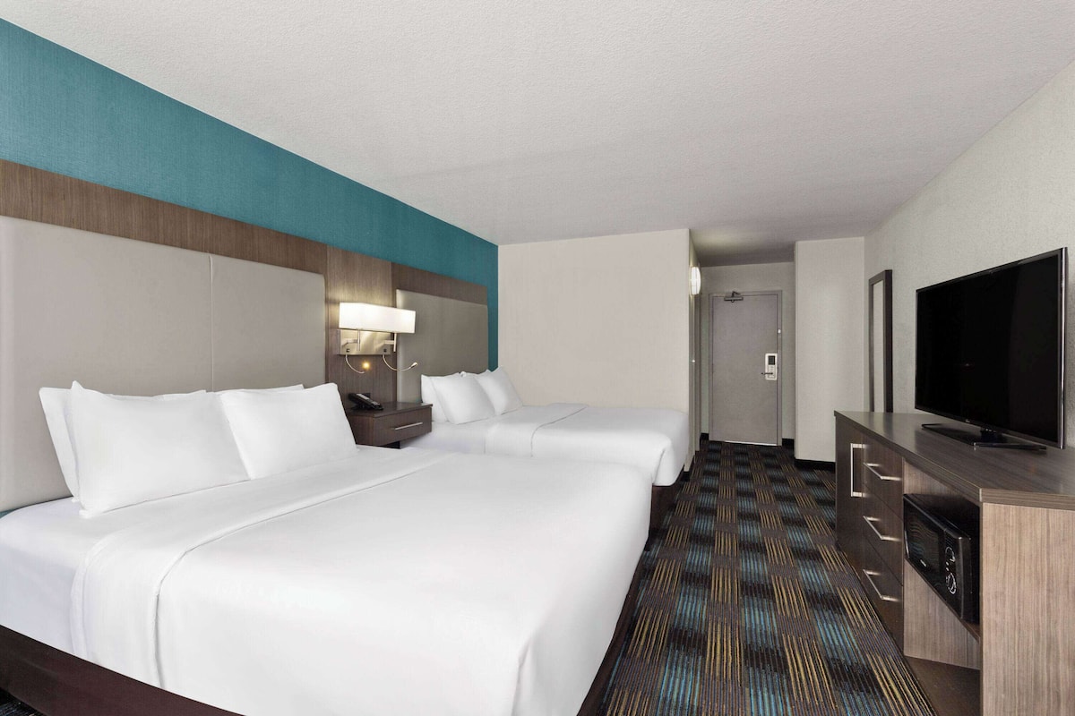 Ein Hotelzimmer mit zwei Betten, einem Fernseher, einem Schreibtisch und einem Badezimmer im Hintergrund.