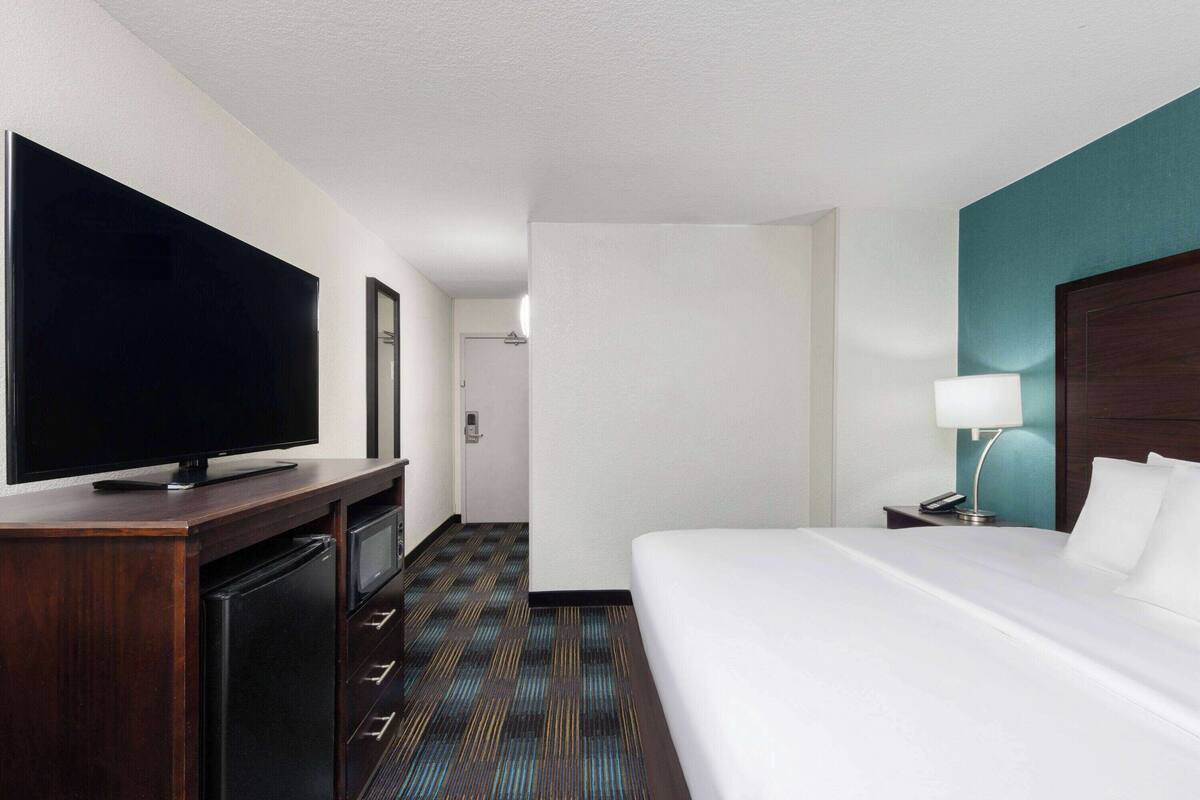 Ein Hotelzimmer mit großem Flachbildfernseher, einem Bett mit weißer Bettwäsche, einem Nachttisch mit Lampe und Teppichboden.