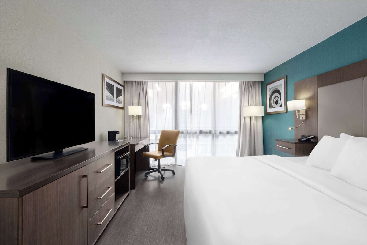 Ein Hotelzimmer mit einem großen Bett, einem Schreibtisch mit Flachbildfernseher, einem Sessel und einem Nachttisch mit Lampe.