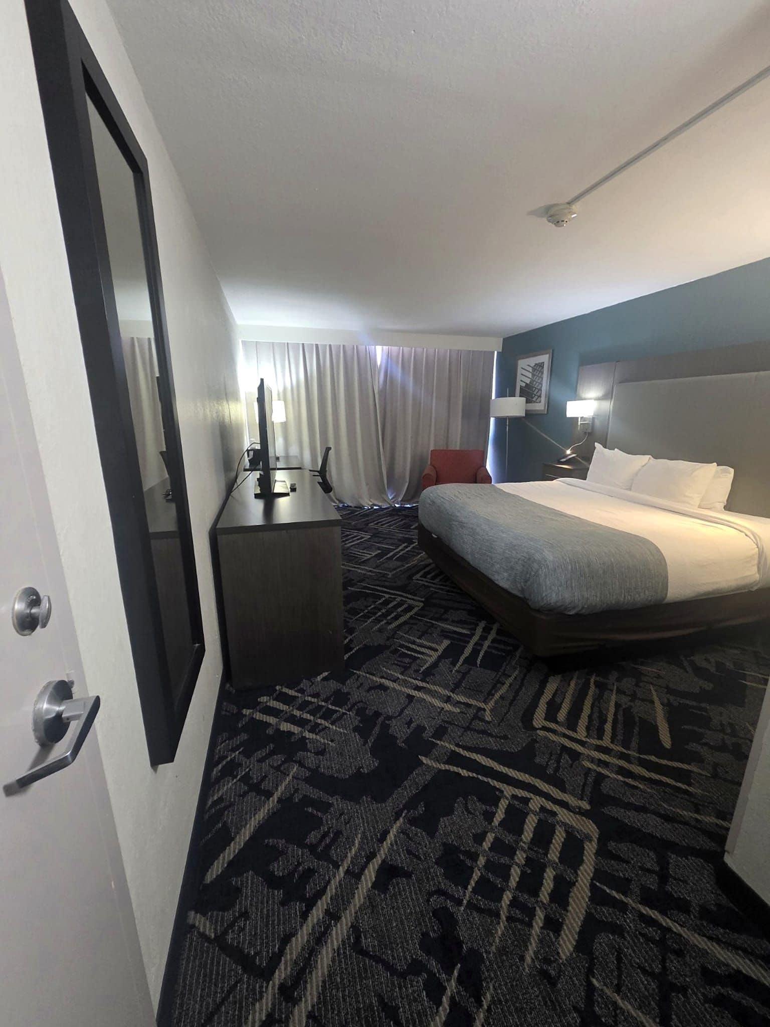 Een hotelkamer met een bed, een bureau, een stoel, een groot raam met gordijnen, en een kamerbreed tapijt met een patroon.