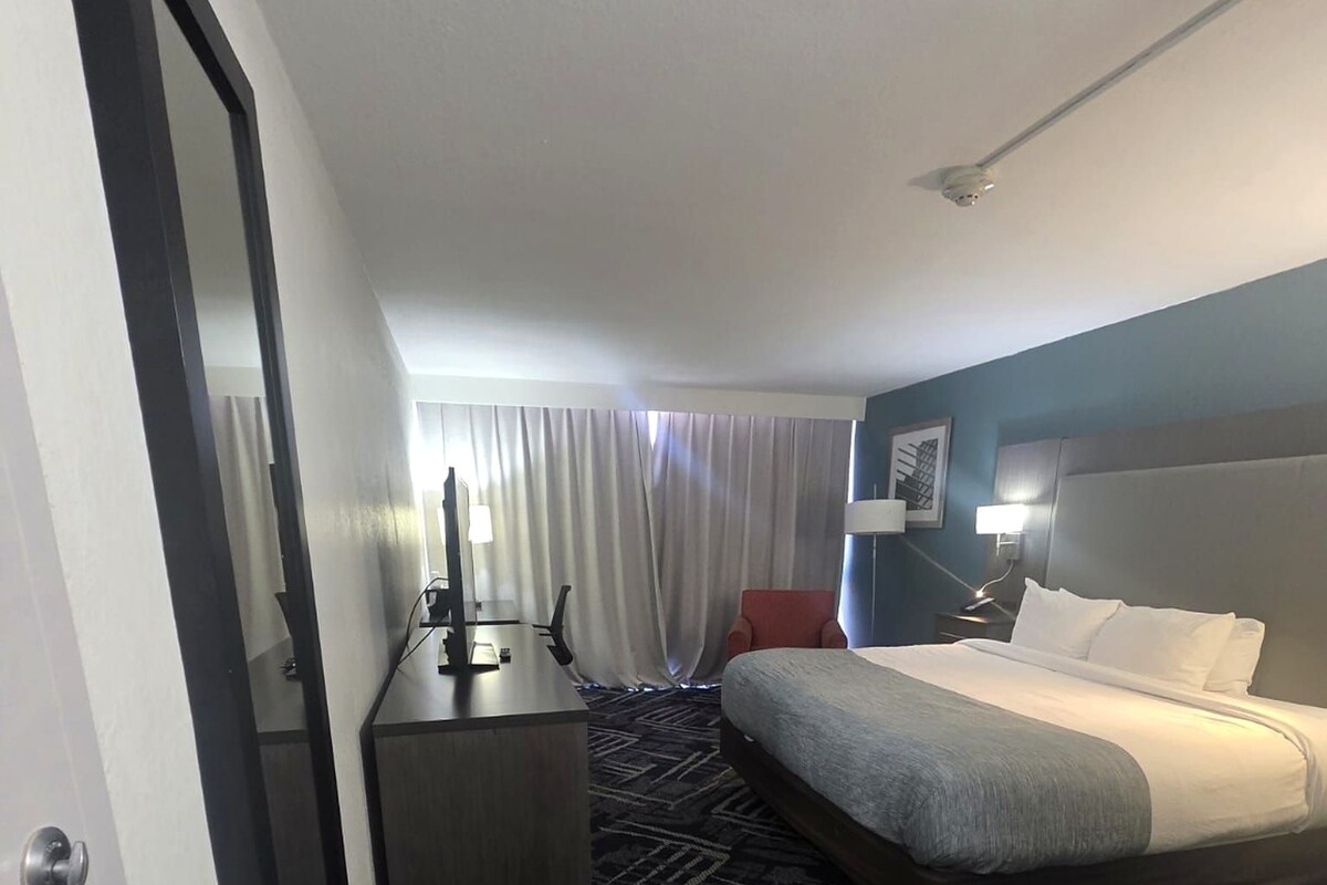 Hotellihuone, jossa on sänky, työpöytä, tuoli, suuri ikkuna verhoilla ja matolla peitetty lattia.