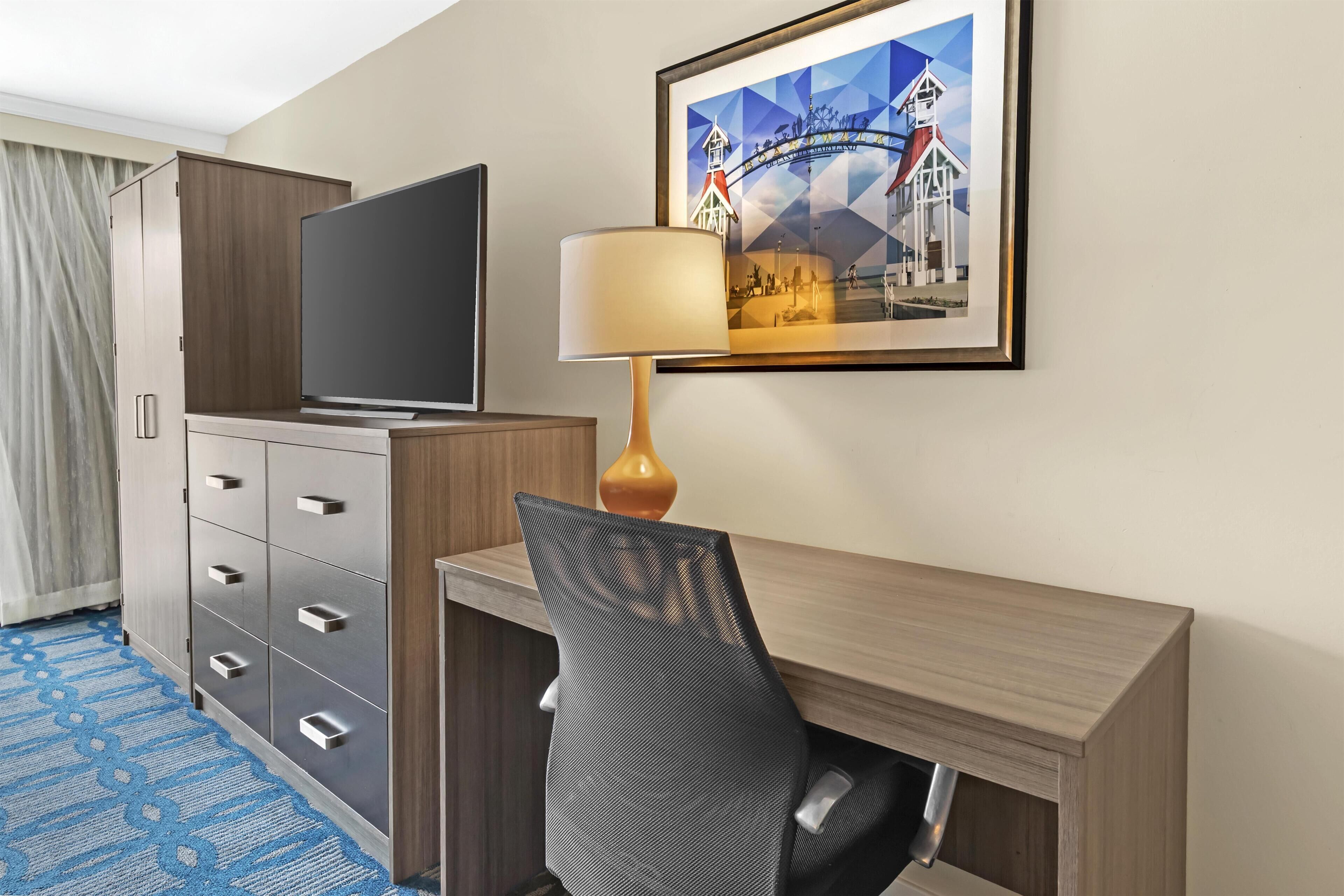 Quarto de hotel com televisão, escrivaninha, cadeira, luminária e quadro na parede.
