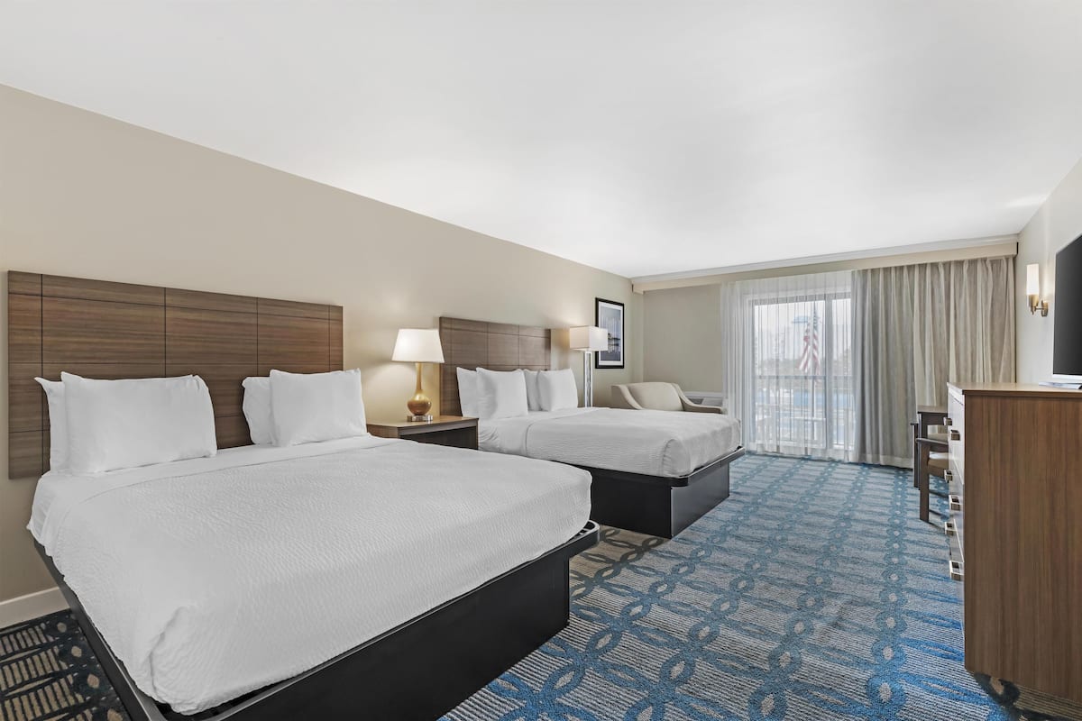 A hotel room with two beds, a desk, a chair, a lamp, and a large window with curtains.