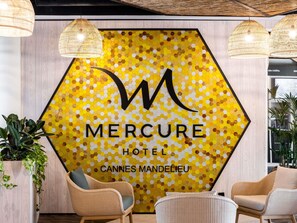 A hotel reception area with a large hexagonal sign displaying the hotel name 'Mercure' and location 'Cannes Mandelieu.' There are wicker chairs and a potted plant in the foreground.