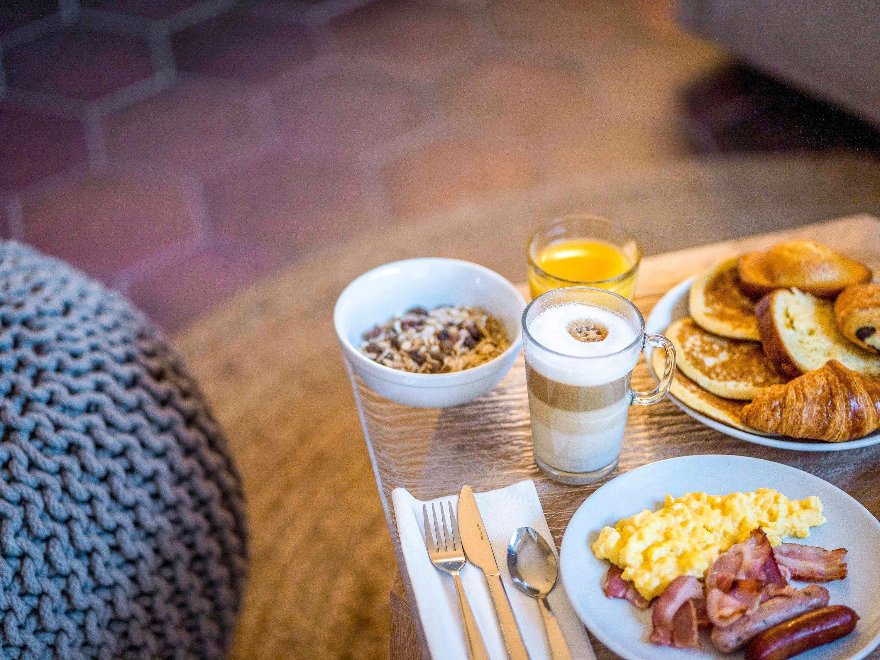 Una mesa de desayuno con un tazón de cereal, un vaso de jugo de naranja, panqueques, croissants, huevos revueltos y tocino.