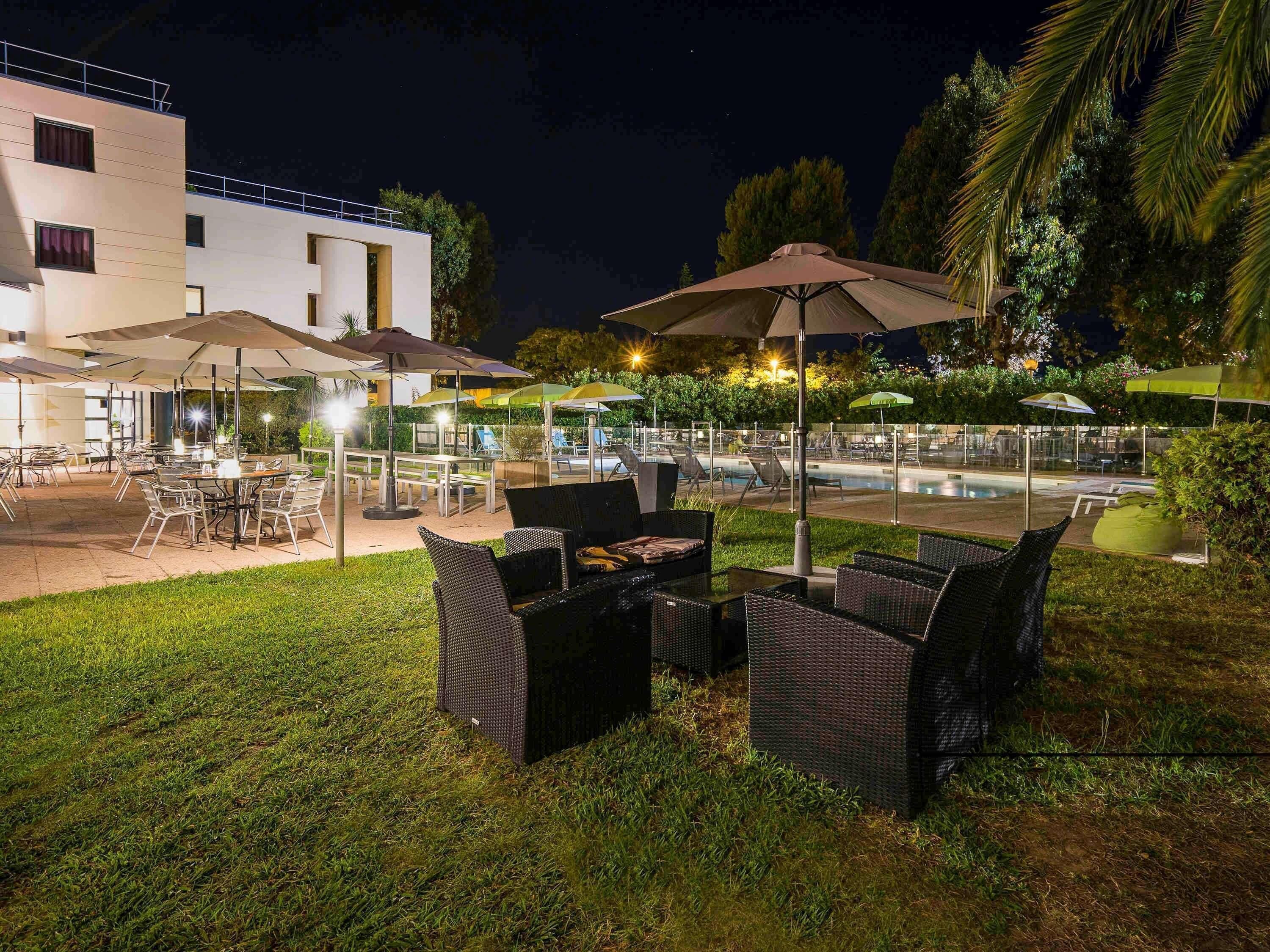 An outdoor dining area with tables and chairs, umbrellas, and a pool in the background.