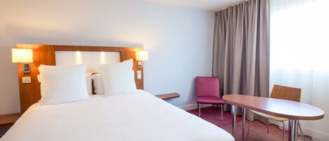 Una habitación de hotel con una cama grande, una mesita pequeña, una silla y un ventanal con cortinas.