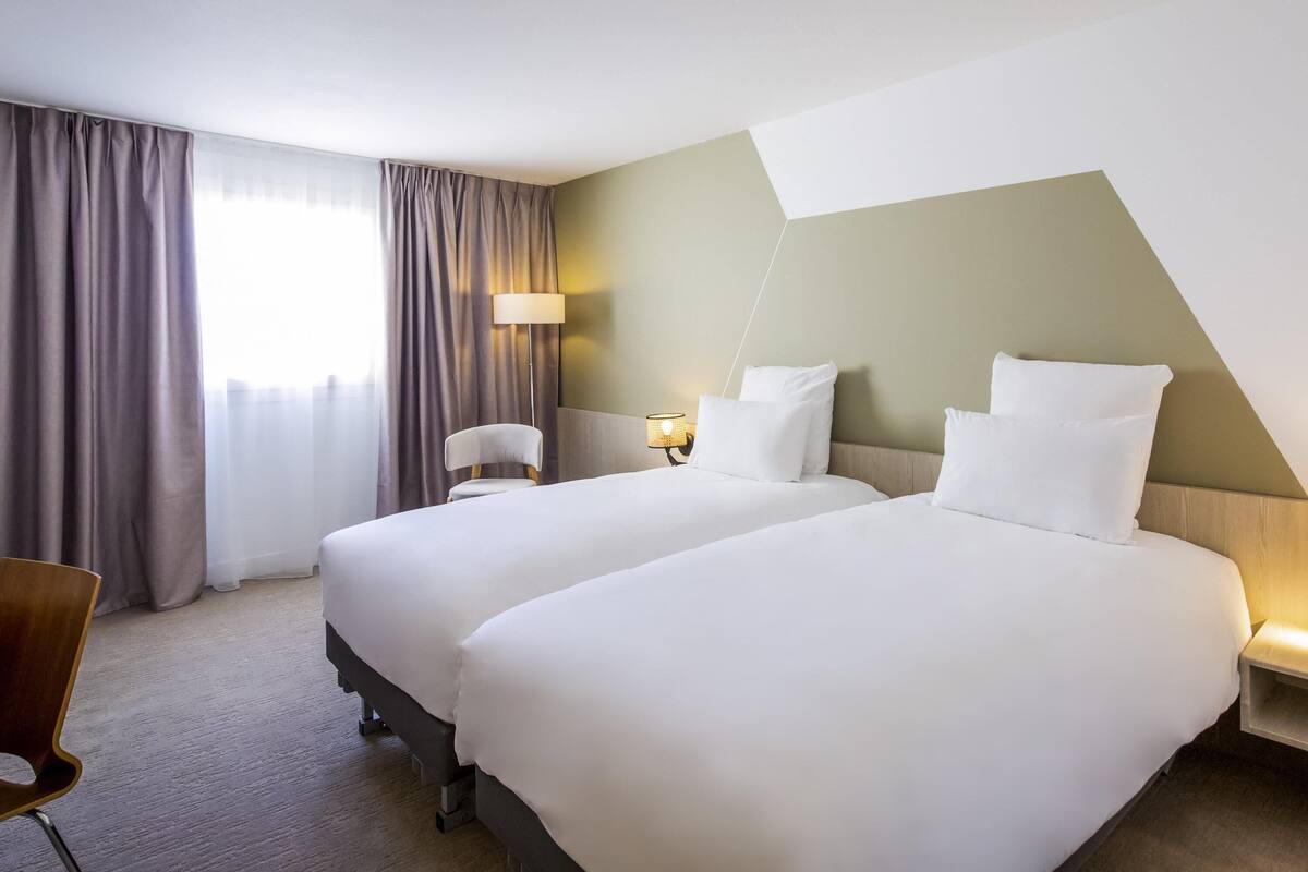 A hotel room with two beds, a chair, a lamp, and a window with curtains.