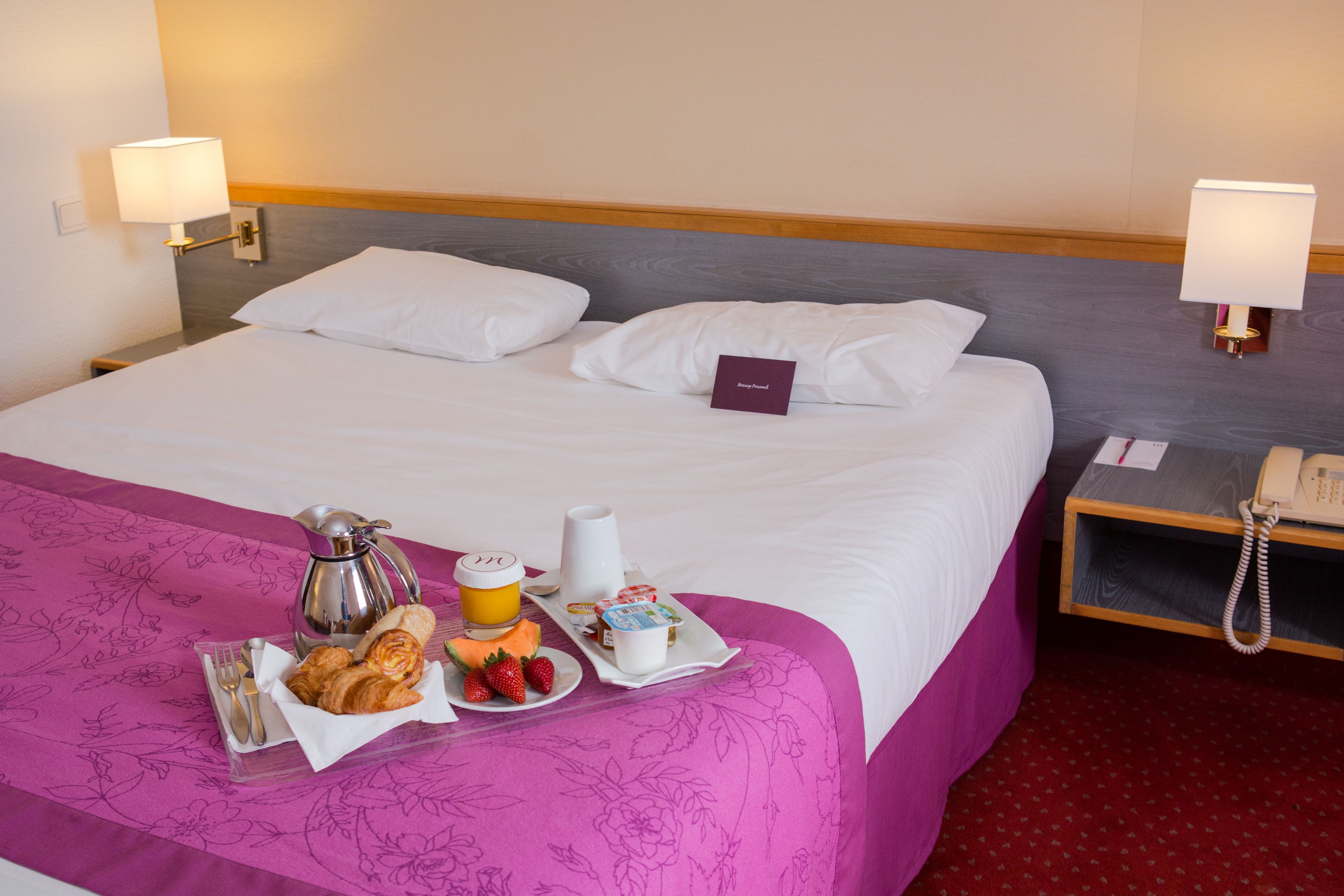 Habitación de hotel con una cama, una mesita de noche con una lámpara, un teléfono y una bandeja con alimentos para el desayuno, incluyendo croissants, fresas y zumo de naranja.