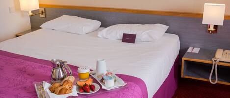 Habitación de hotel con una cama, una mesita de noche con una lámpara, un teléfono y una bandeja con alimentos para el desayuno, incluyendo croissants, fresas y zumo de naranja.