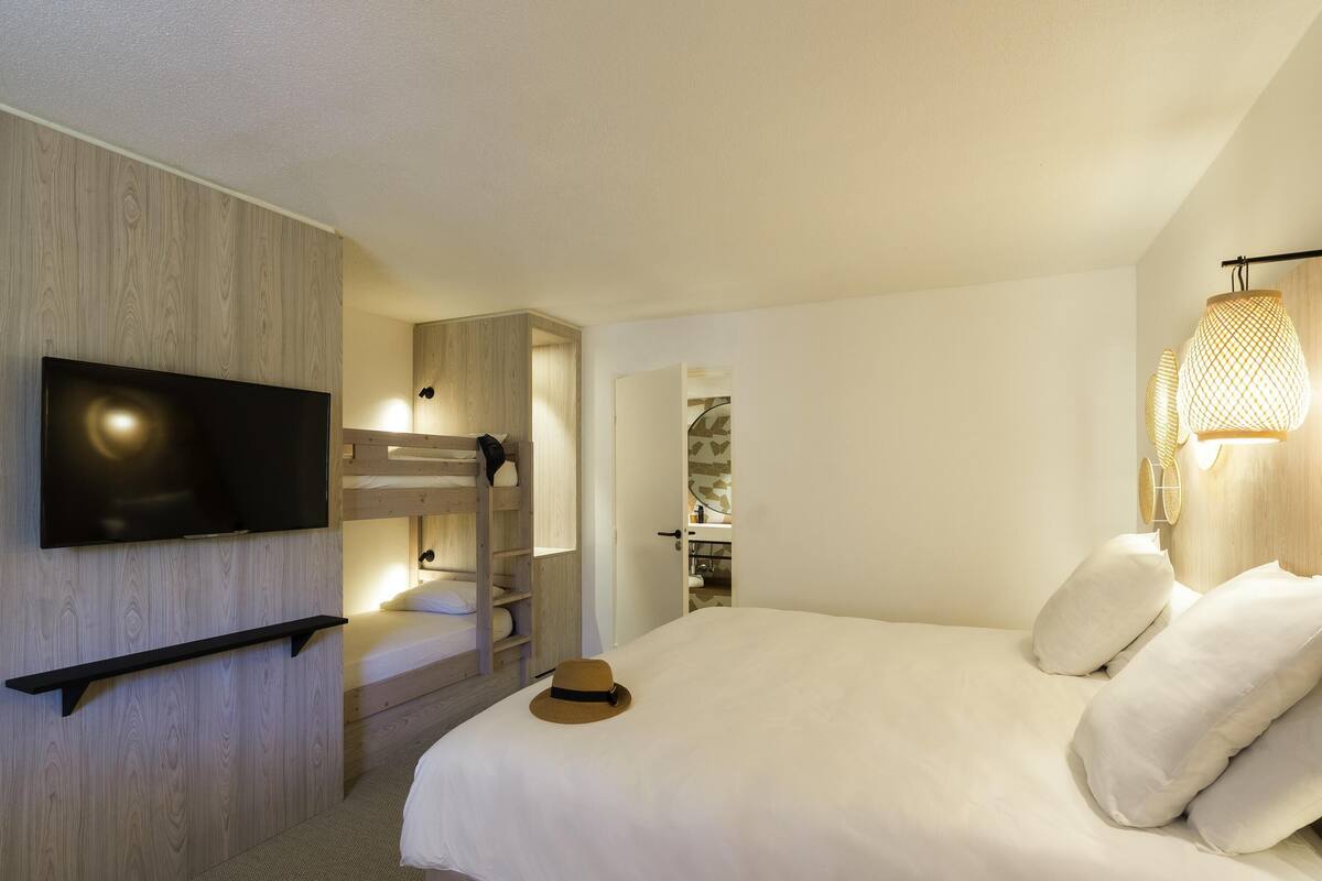 A modern hotel room with a bunk bed, a flat-screen TV mounted on the wall, a bedside table, and a lamp.
