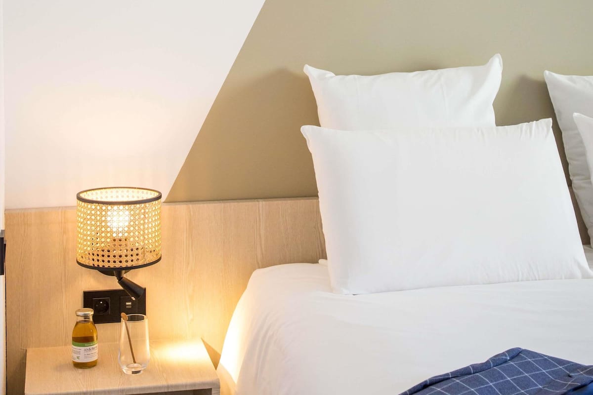 A hotel room with a bed, white pillows, a nightstand with a lamp, a bottle, and a glass.