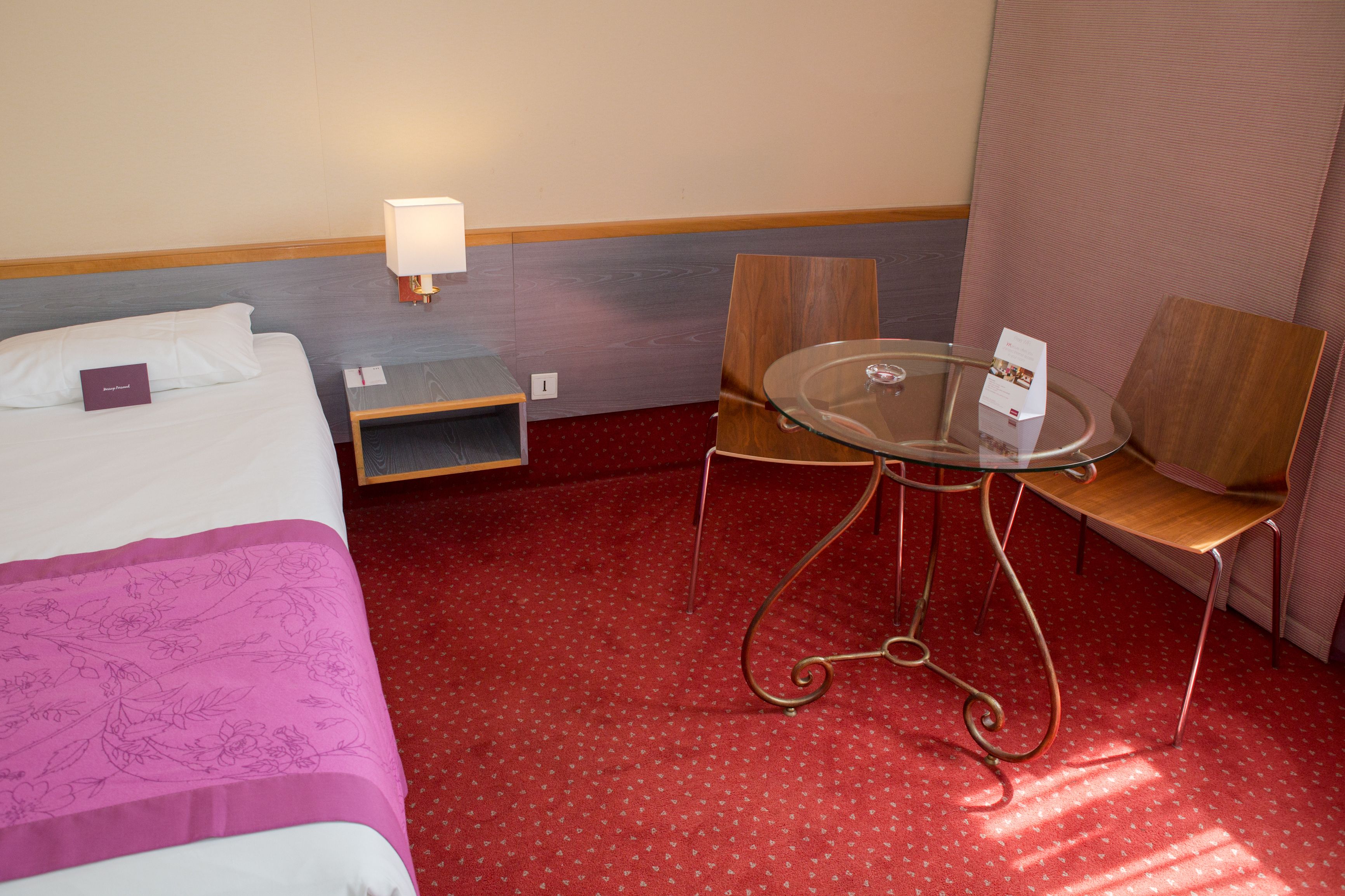A hotel room with a bed, a small table with chairs, a lamp, and a red carpeted floor.
