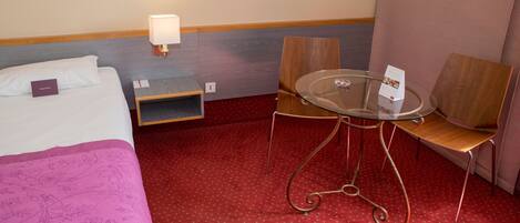 A hotel room with a bed, a small table with chairs, a lamp, and a red carpeted floor.