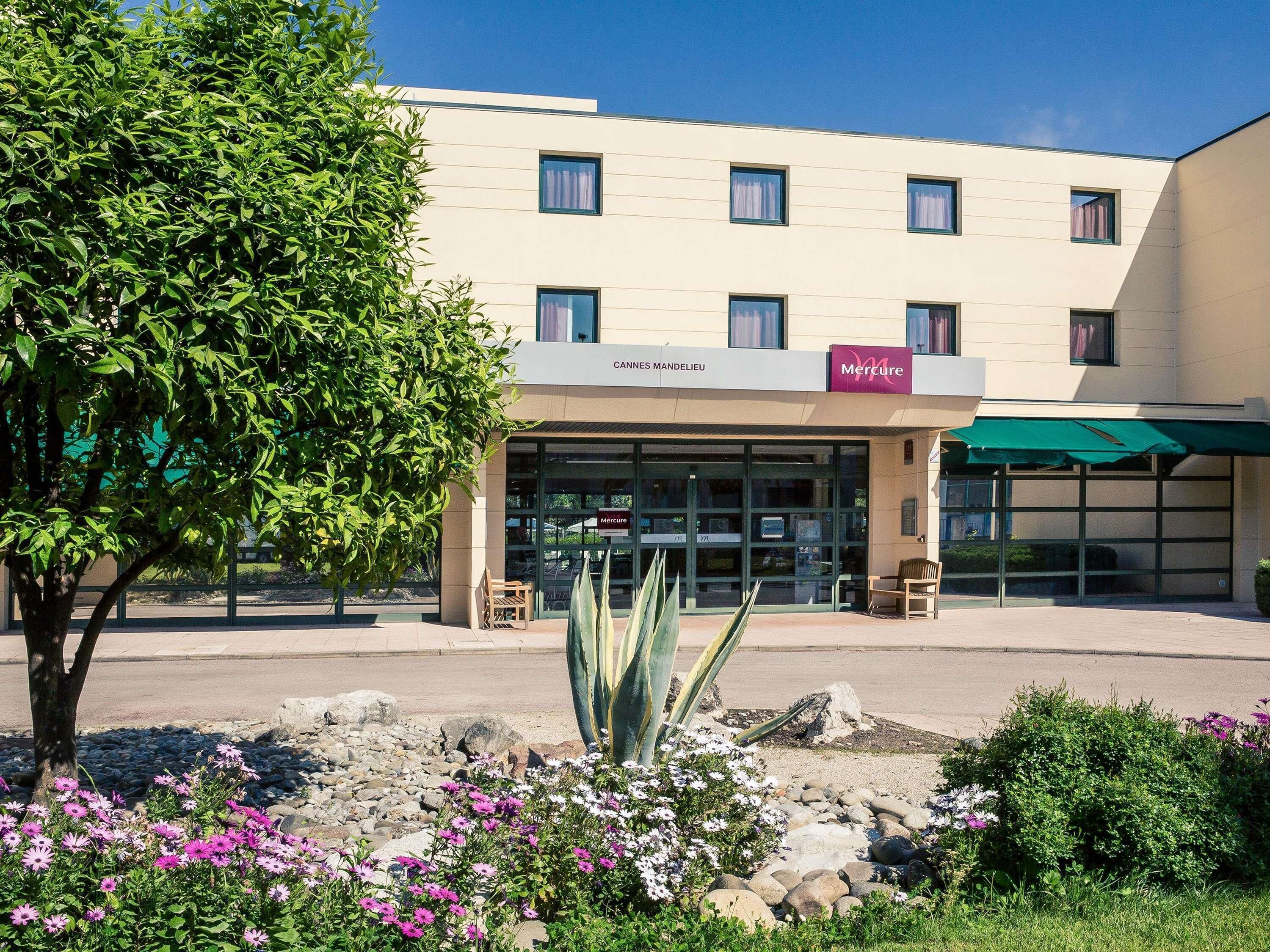 A building with a sign for 'Mercure' and 'Cannes Mandelieu' hotel. There are benches outside, a tree, and a garden with rocks and flowers.