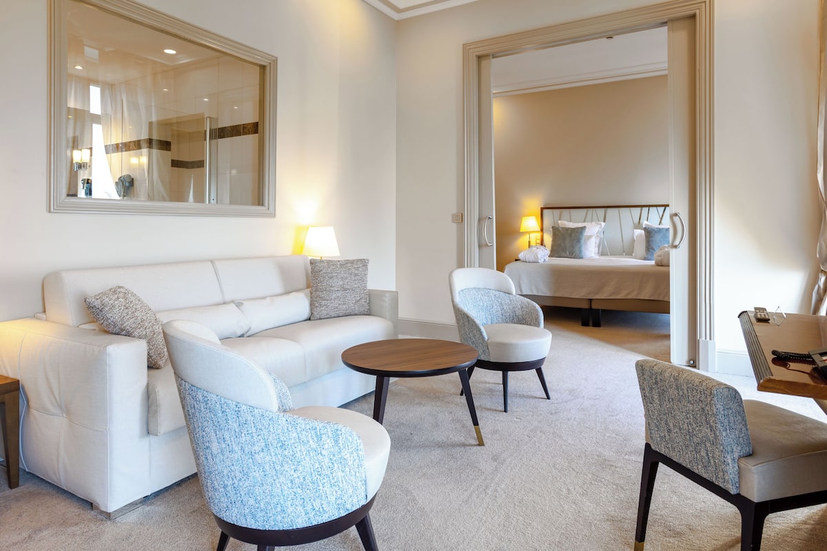 Moderni hotellihuone, jossa on sohva, kaksi nojatuolia, sohvapöytä, sänky ja työpöytä sekä kitara.