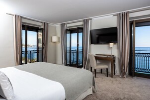 Habitación de hotel con cama, escritorio, silla, televisor y balcón con vista al mar.