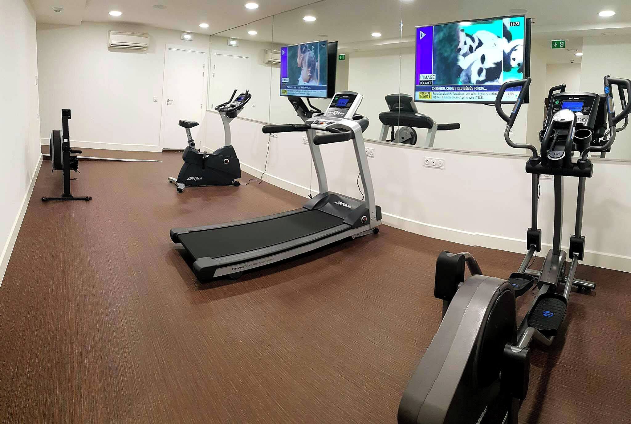 Un gimnasio con cintas de correr, elípticas y una pantalla de televisión mostrando un programa.