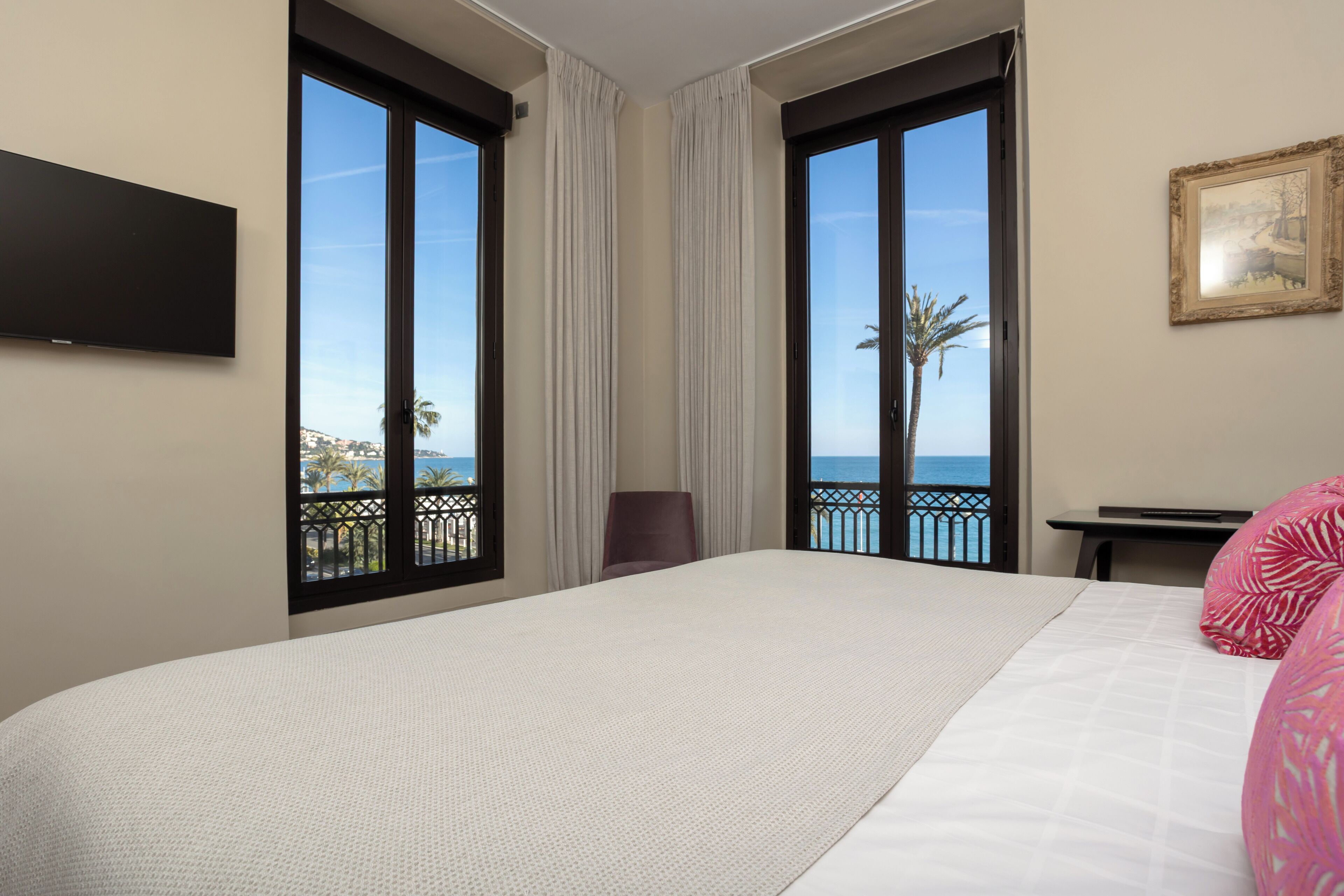 Habitación de hotel con una cama grande, un escritorio, una silla y un televisor. Hay dos ventanas amplias con cortinas y vistas al mar y palmeras.