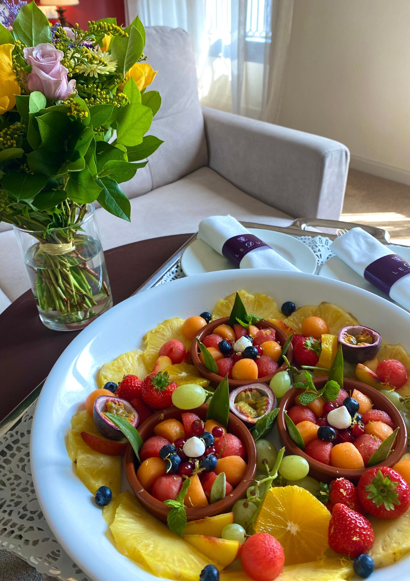 A dining table with a white plate of fruit salad and a vase of flowers.