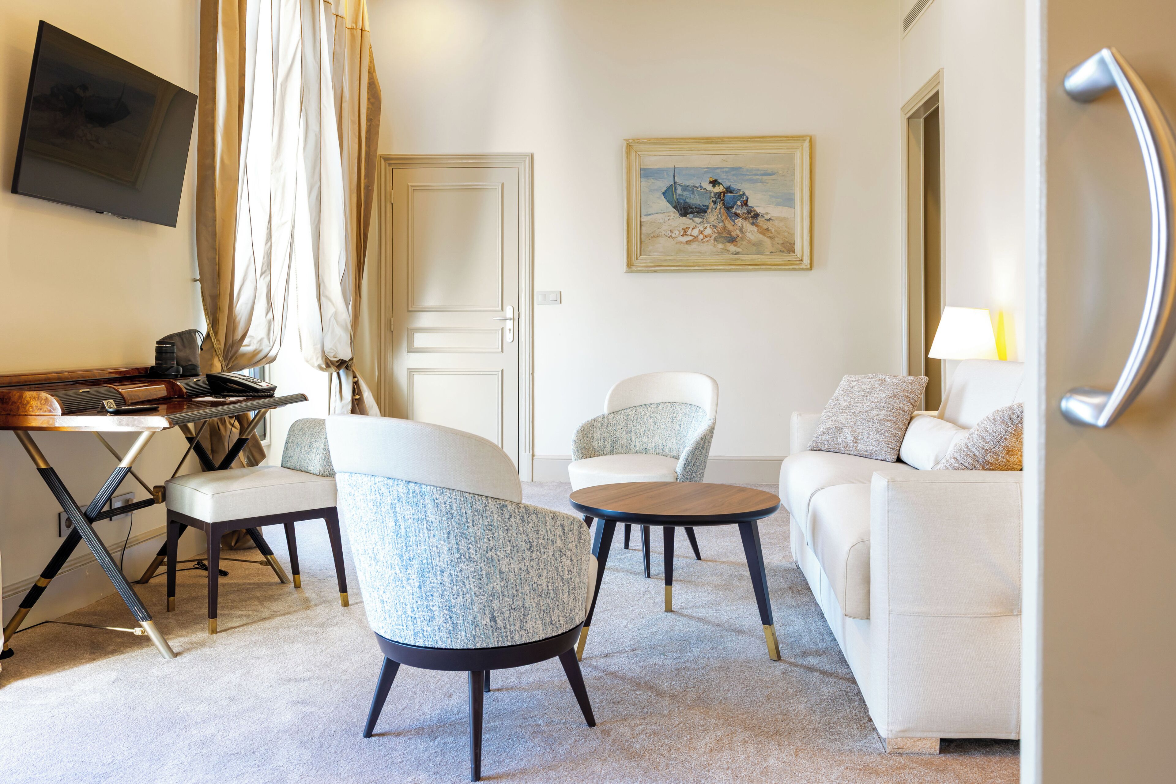 A hotel room with a white sofa, two armchairs, a small table, a flat-screen TV mounted on the wall, and a painting on the wall.