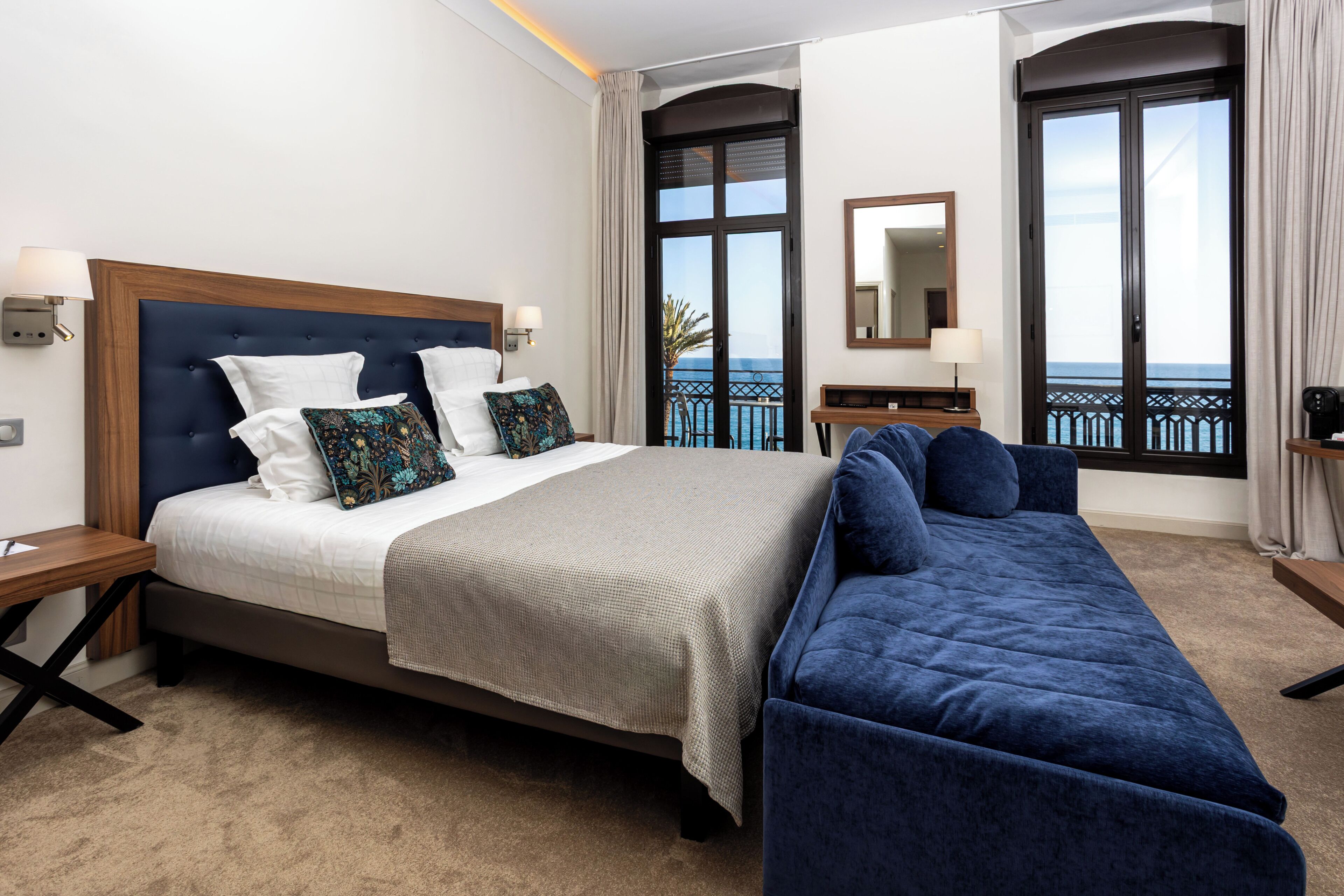 A hotel room with a bed, a sofa, a nightstand, a lamp, a mirror, and a view of the sea through large windows.