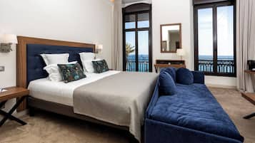 A hotel room with a bed, a sofa, a nightstand, a lamp, a mirror, and a view of the sea through large windows.