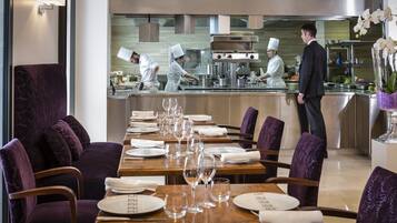 Un restaurante con una larga mesa de comedor dispuesta para una comida, una zona de cocina con cocineros preparando alimentos y un camarero junto al mostrador.