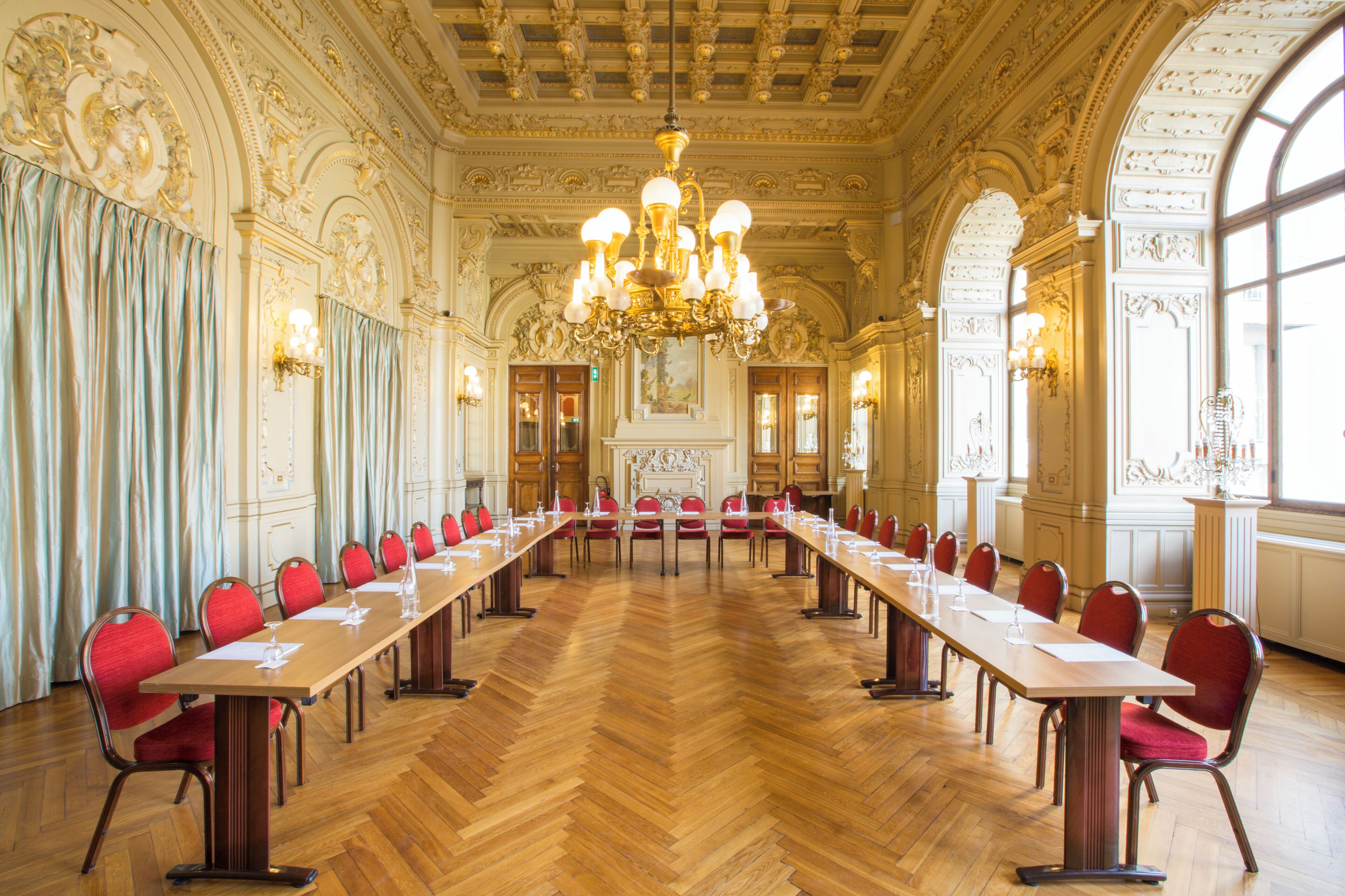 Una larga mesa de conferencias con sillas, techo ornamentado, candelabros y ventanas grandes.