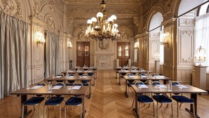 A grand conference room with ornate ceiling and wall decorations, multiple tables with chairs, and chandeliers.