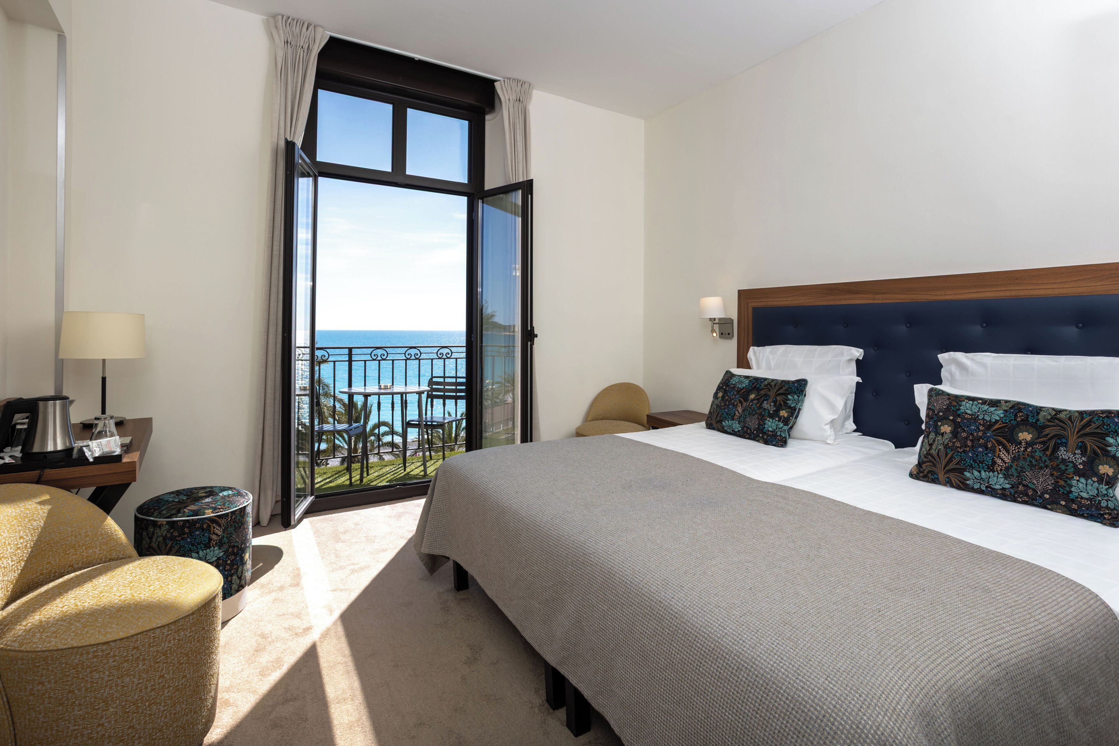 A hotel room with a large bed, a desk with a lamp, a chair, and a view of the sea through a balcony.