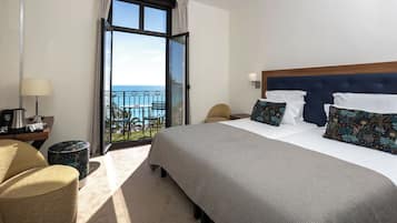 A hotel room with a large bed, a desk with a lamp, a chair, and a view of the sea through a balcony.