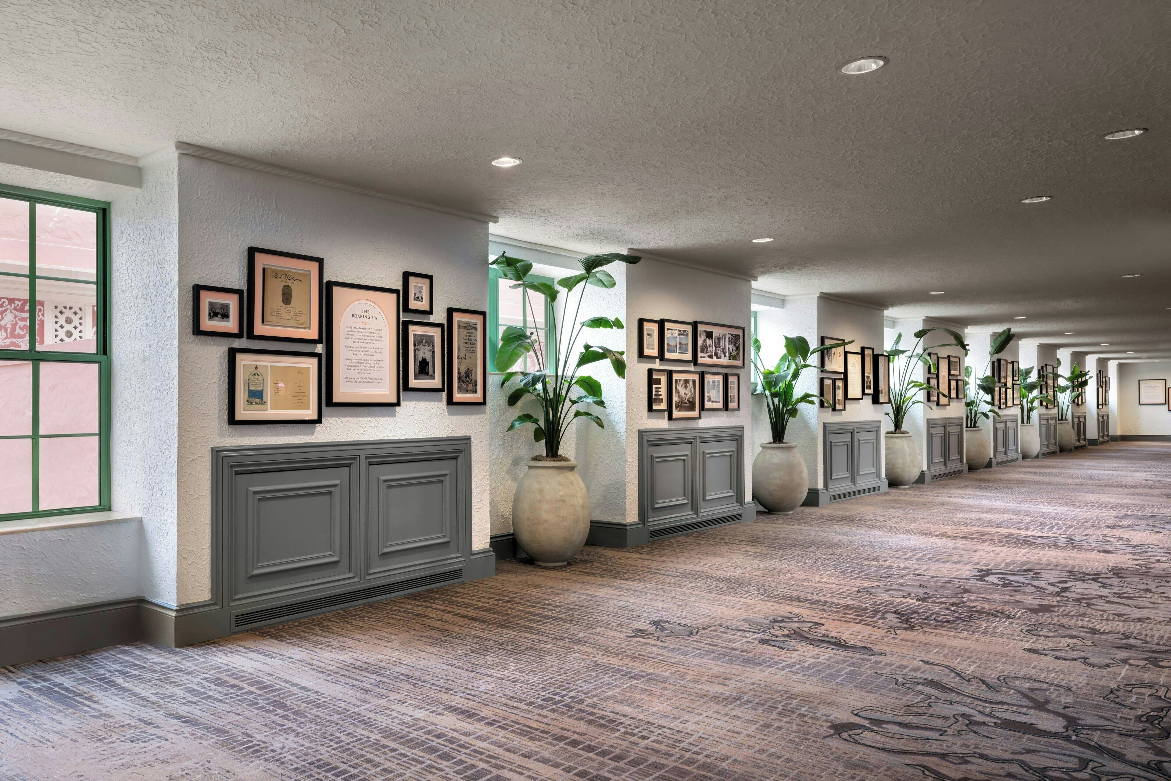 A long hallway with framed pictures on the walls, large potted plants, and a patterned carpet.