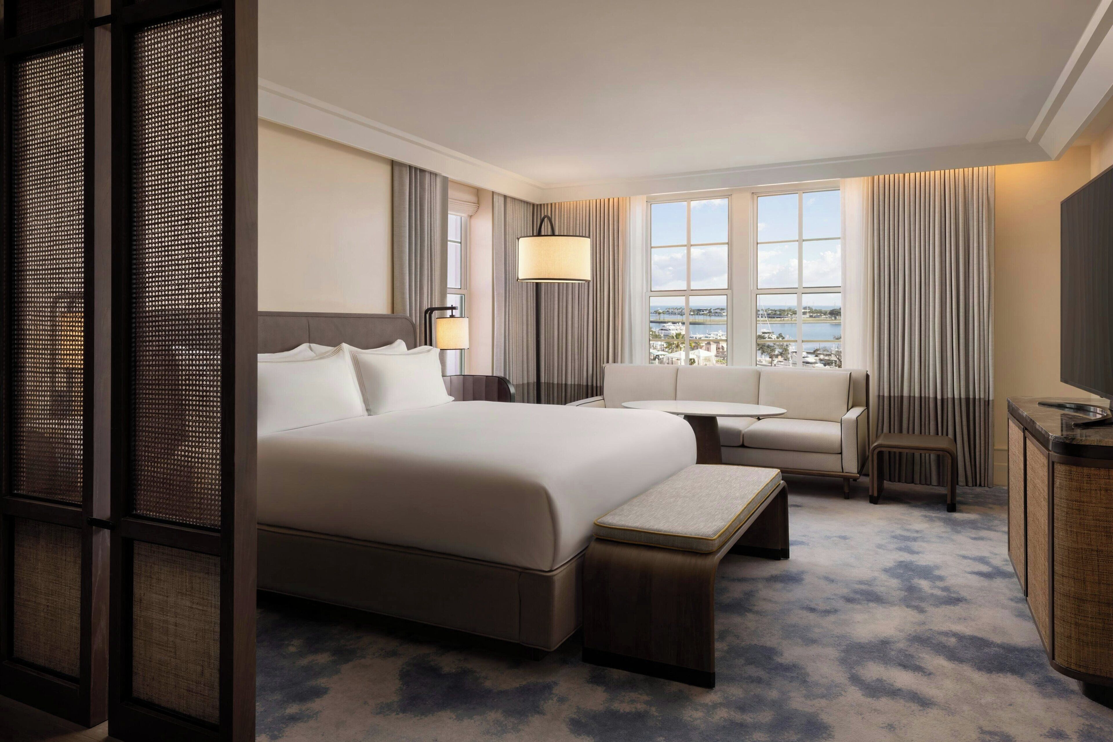 A hotel room with a large bed, a sofa, a small table, a TV, and a view of the city.
